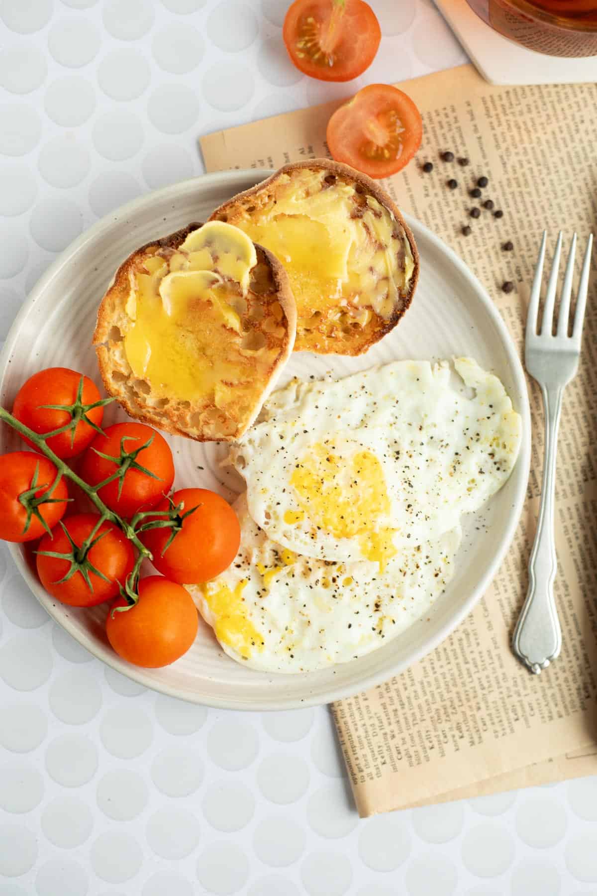 2 over hard eggs on a white plate with a buttered engligh muffin and tomatoes on the vine.