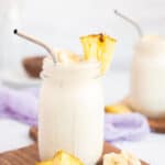 a pineapple banana smoothie in a jar with a chunk of pineapple on the rim with a silver straw.