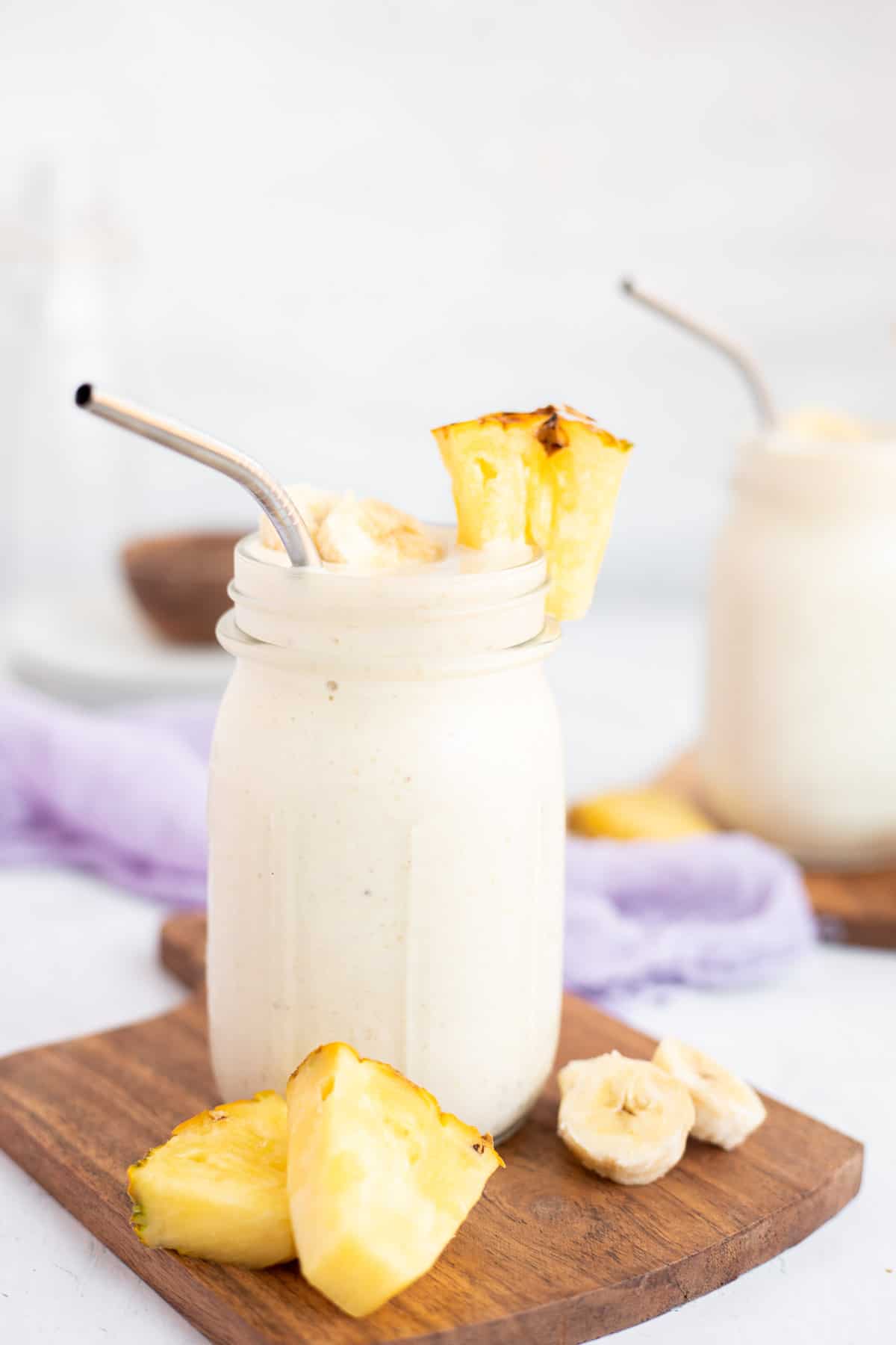 a pineapple banana smoothie in a jar with a chunk of pineapple on the rim with a silver straw.