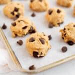 balls of edible protein cookie dough with chocolate chips on a parchment lined baking sheet.
