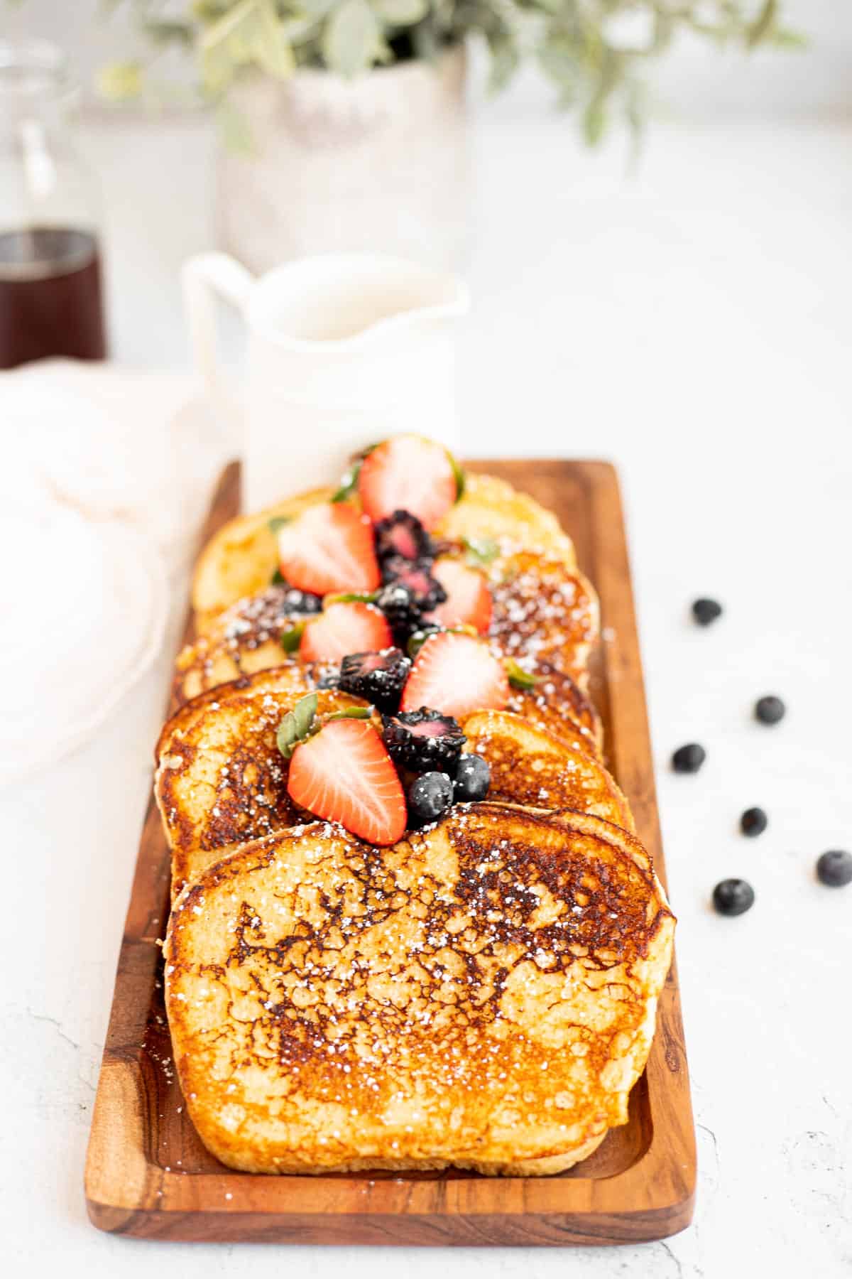 slices of protein French toast lined up on a wooden platter with a white pitcher of syrup.