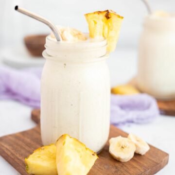 pineapple banana smoothie in a jar with a pineapple chunk on the rim, the jar is on a wooden board.