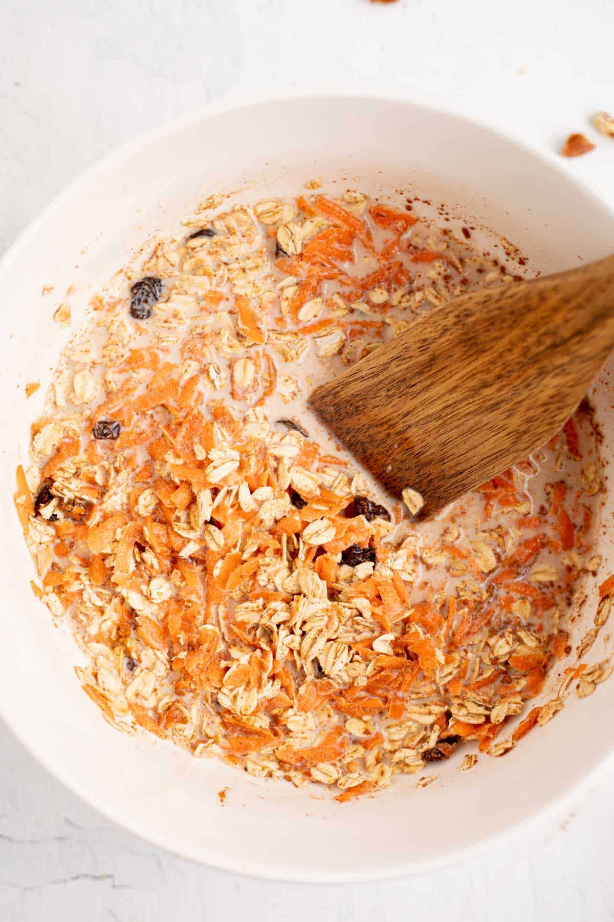 oats, raisins, milk, and shredded carrots in a white bowl with a wooden spoon.