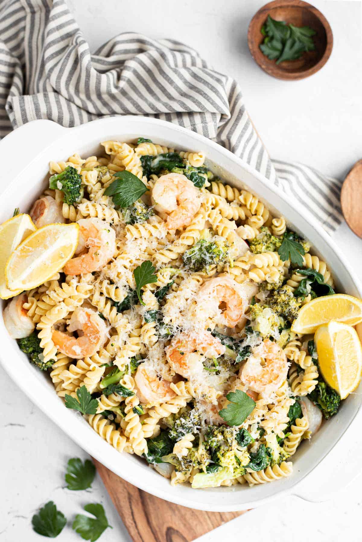 shrimp and broccoli pasta in a white serving dish with lemon wedges and stripped napkin.