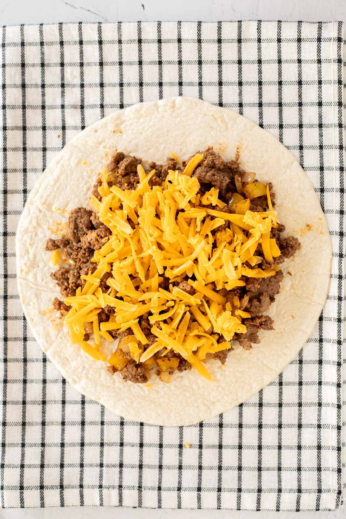 tortilla topped with cooked ground beef and shredded cheese on a checkered tea towel.