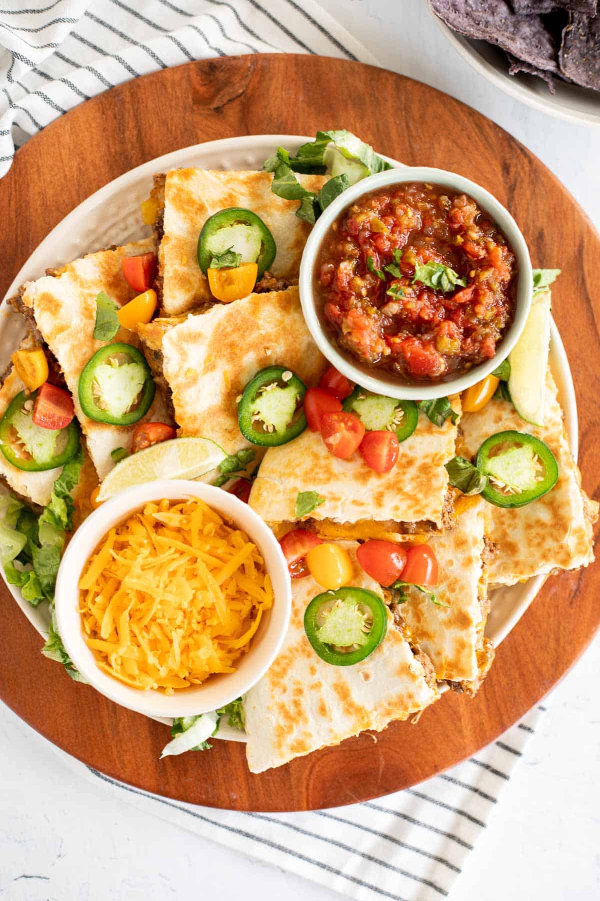 A platter of taco quesadillas with jalapenos, tomatoes, lime wedges, and bowls of salsa and cheese.