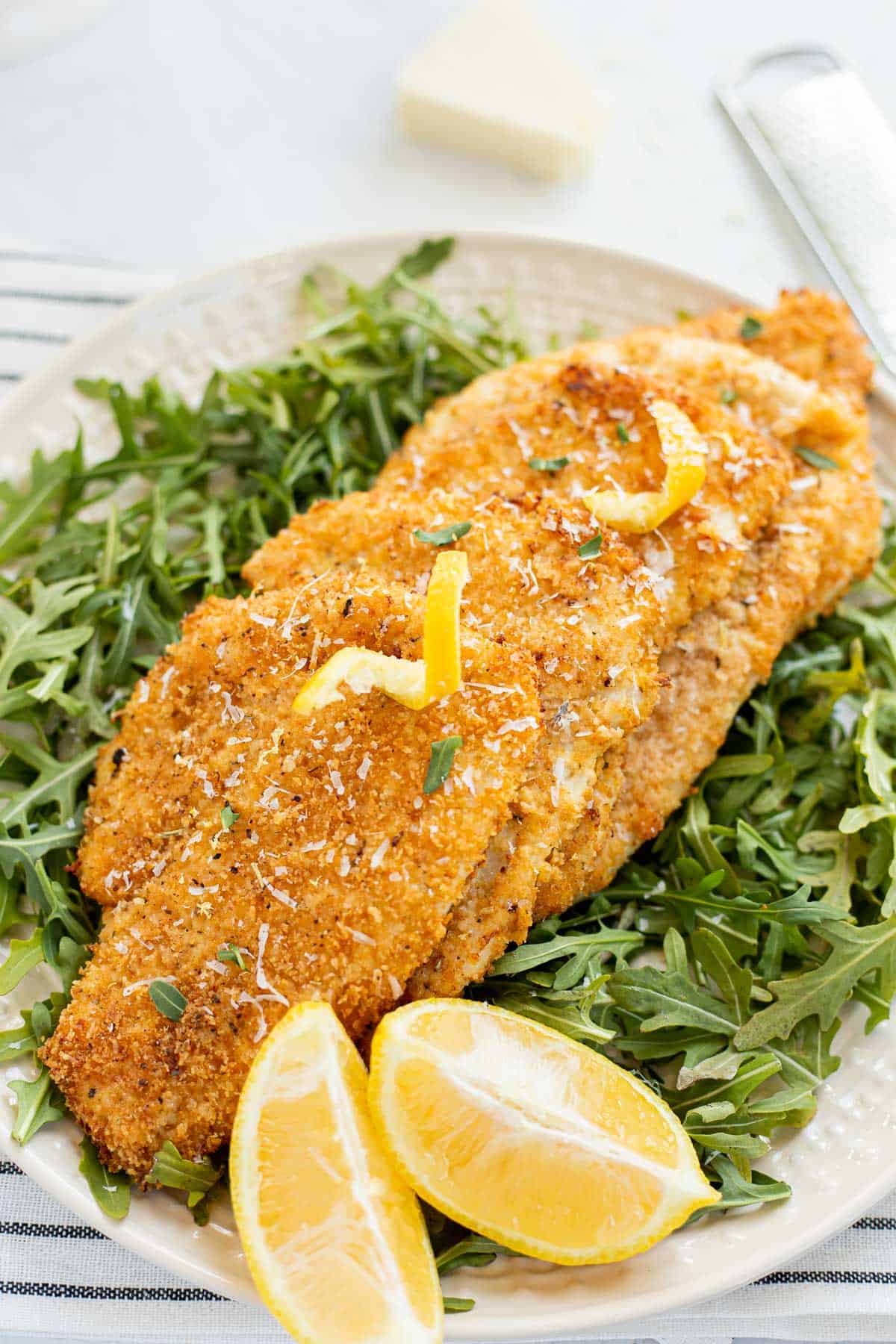 breaded air fryer cutlets on a bed of arugula on a white plate with lemon wedges.