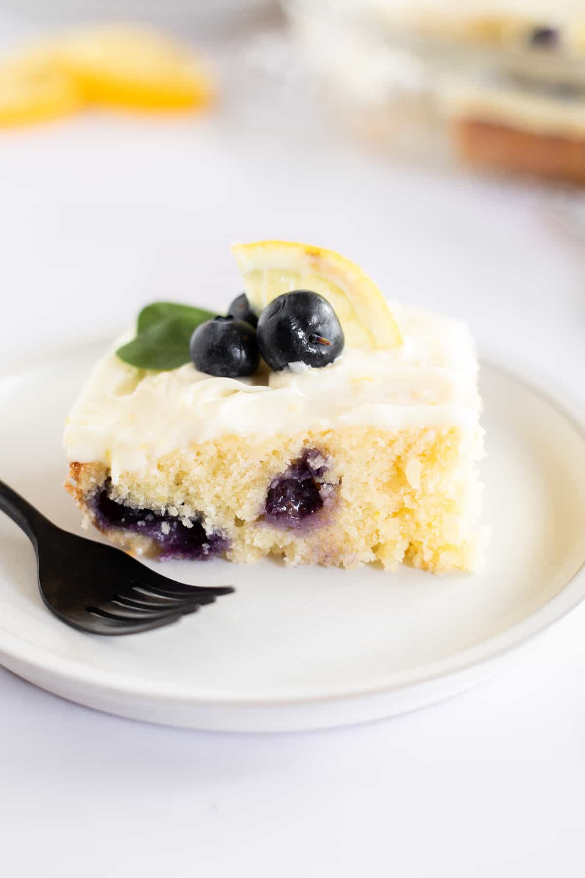 a slice of lemon blueberry sheet cake with cream cheese frosting on a white plate with a black fork.