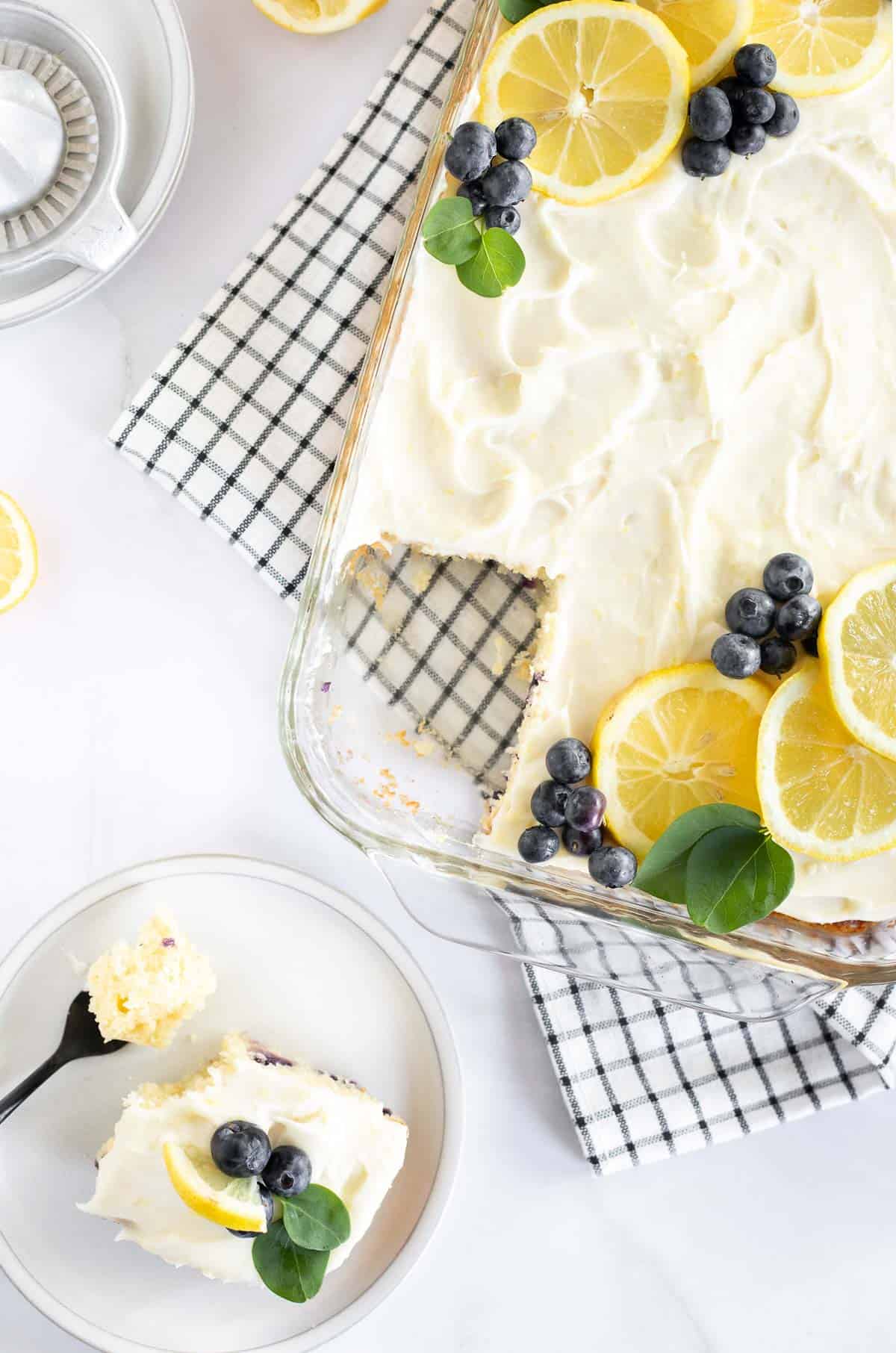 an overhead view of lemon sheet cake with blueberries in a baking dish and a piece on the side.