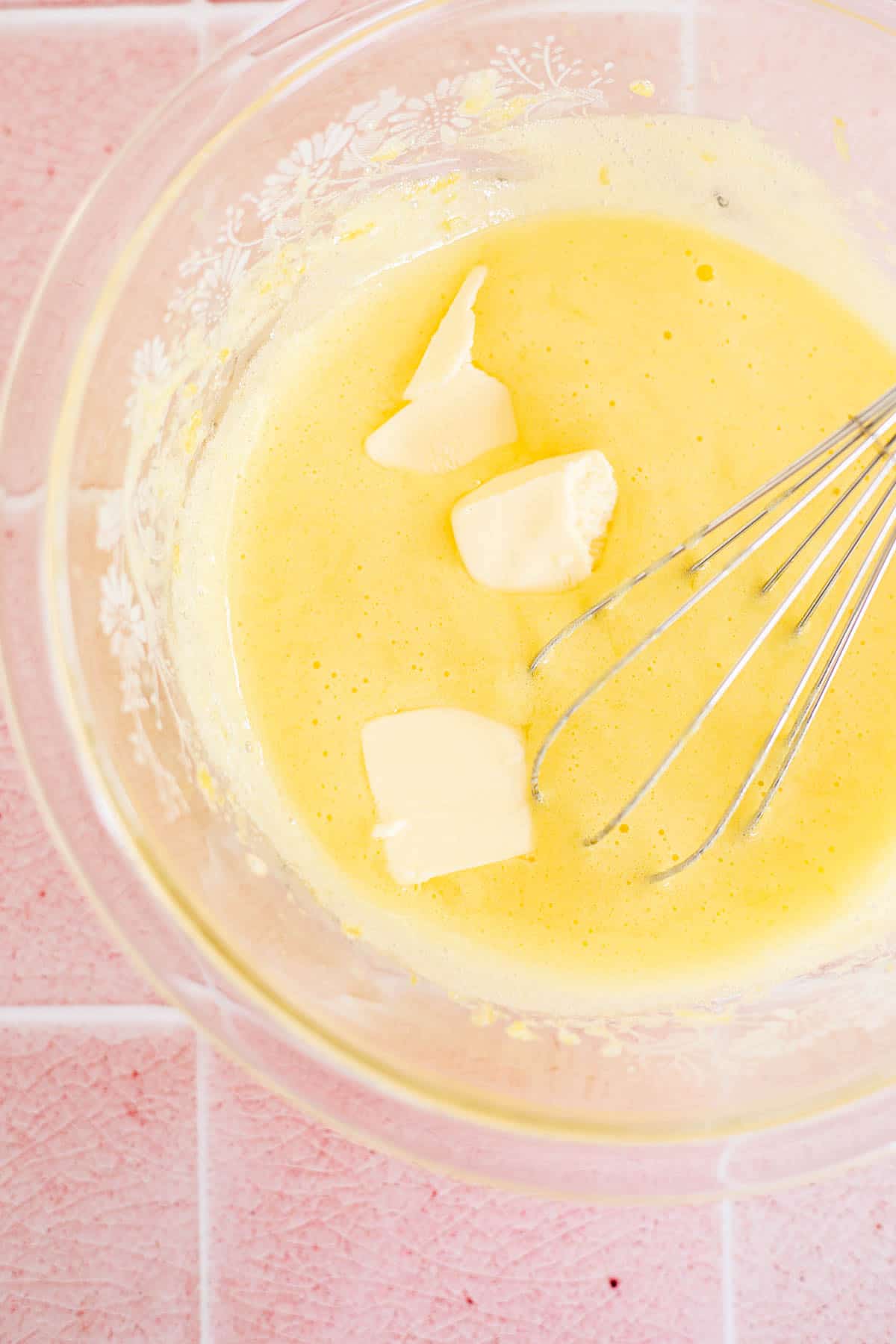 homemade lemon curd with a whisk and pats of butter in a glass bowl.