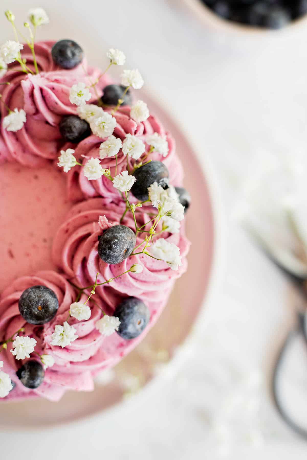 overhead shot of a cake with blueberry buttercream decorated with flowers and fresh berries.