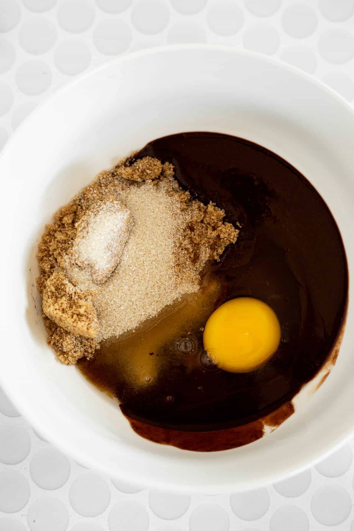 brown sugar, granulated sugar, and an egg in a white bowl of melted chocolate.