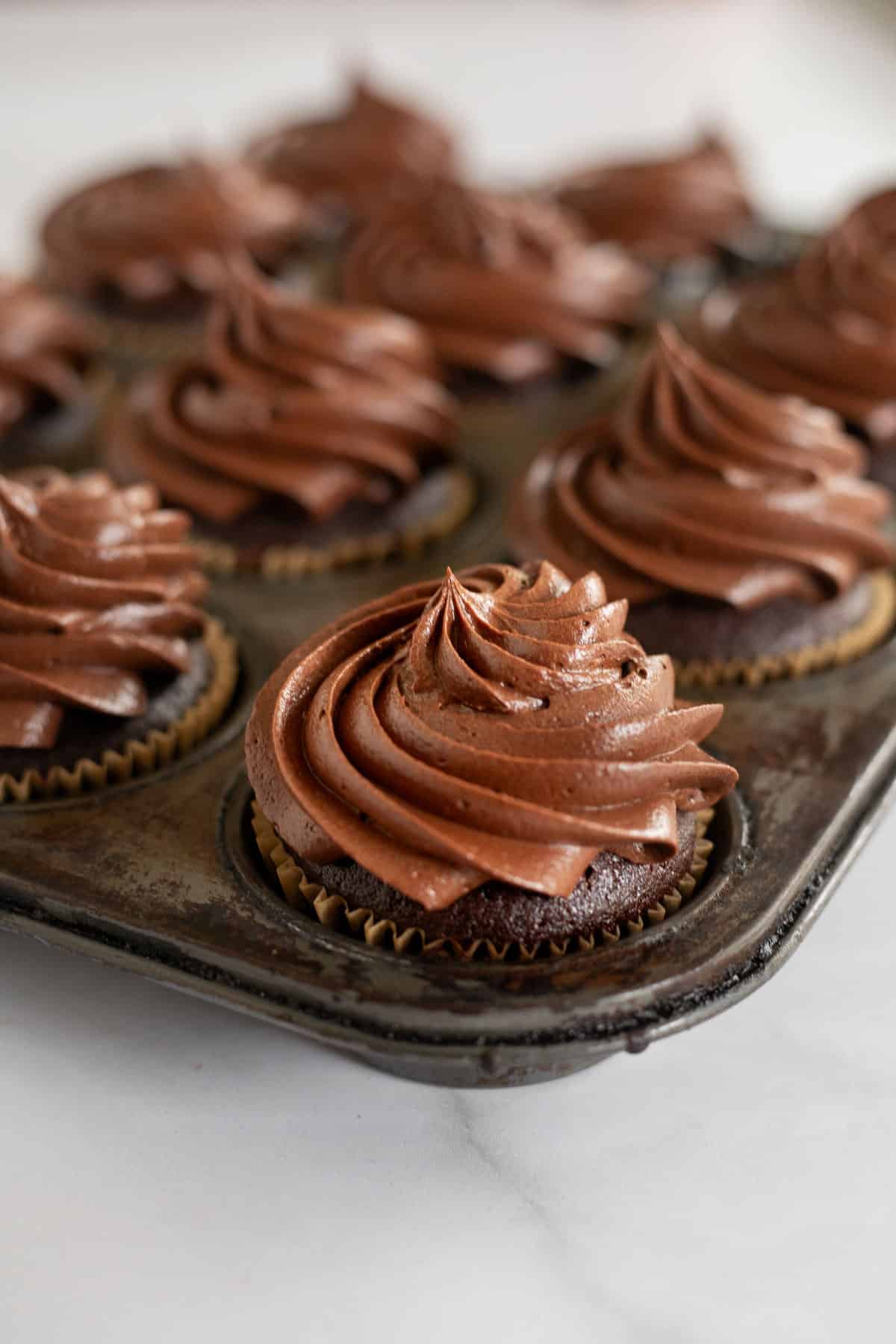 fudgy chocolate ganache frosting piped onto chocolate cupcakes in a muffin pan.