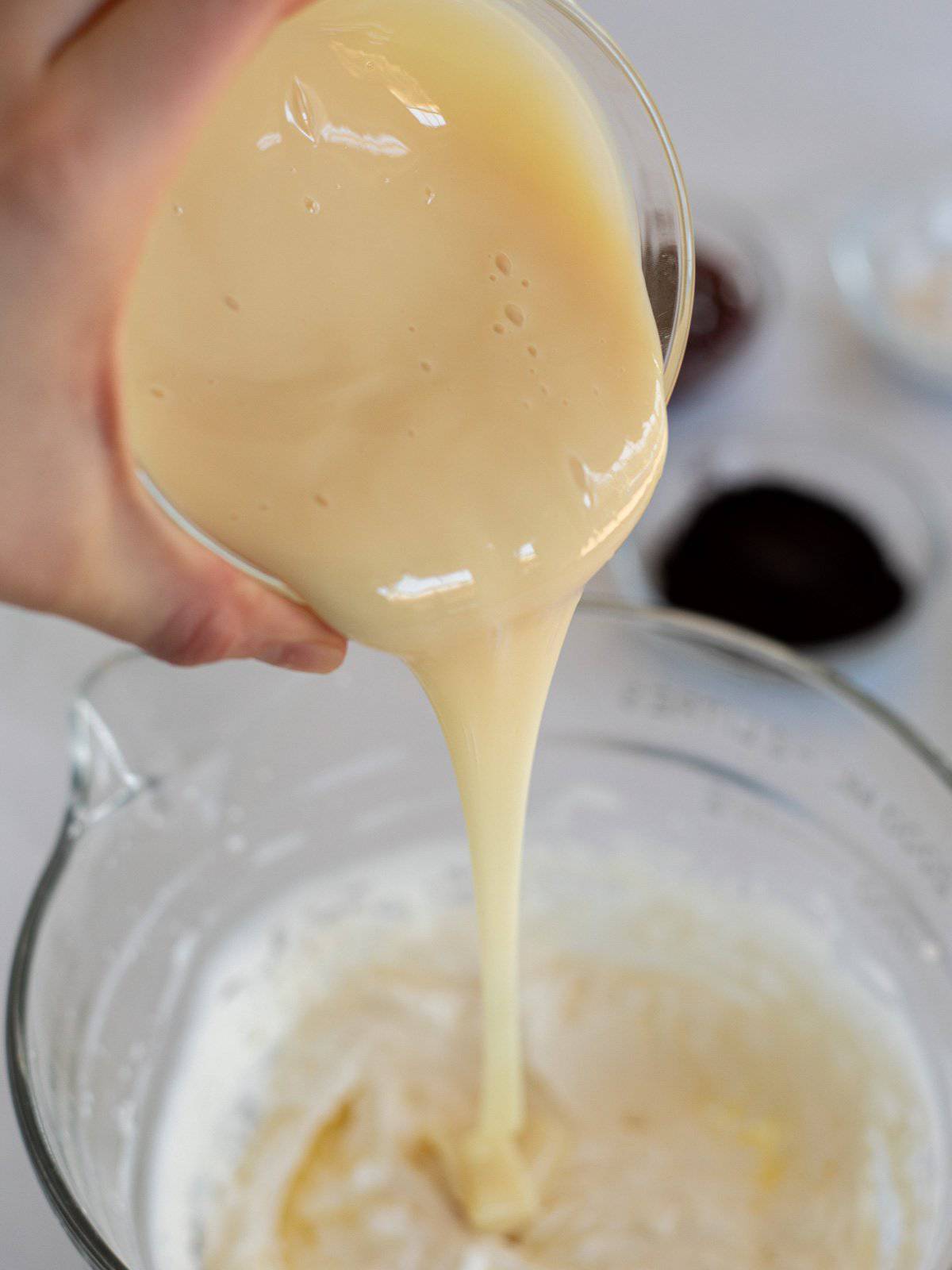 sweetened condensed milk pouring into a glass bowl of whipped cream